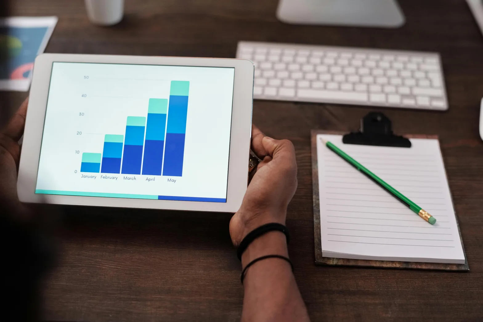 Two hands holding a tablet which displays a blue graph, with a clipboard and keyboard beside it.